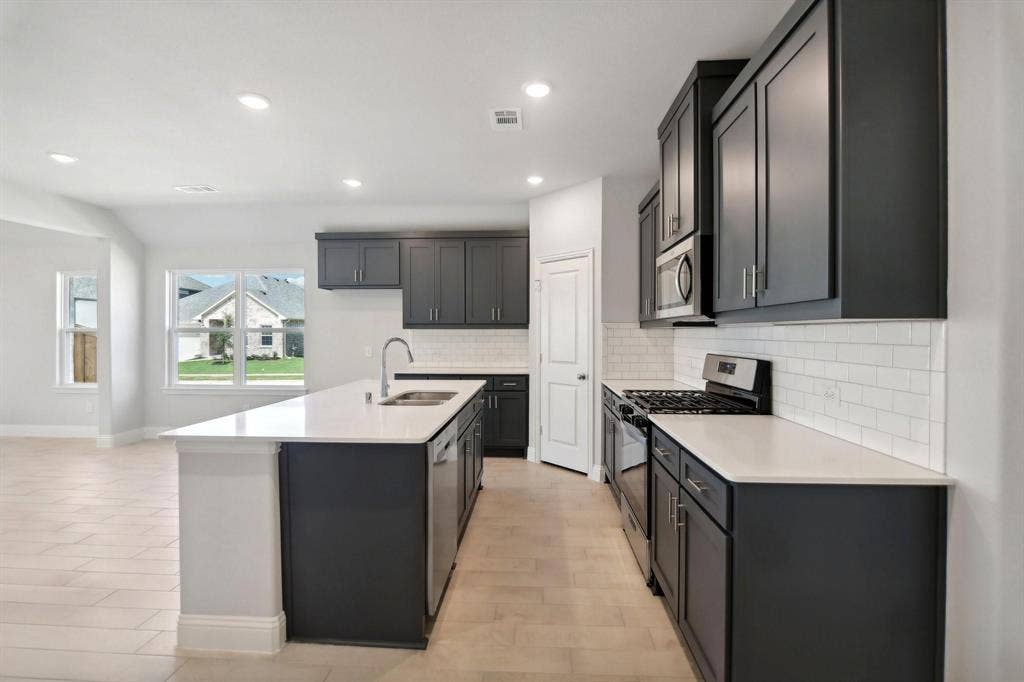 Kitchen featuring an island with an undermount sink, stainless steel appliances, dark cabinetry, light countertops, and a subway tile backsplash