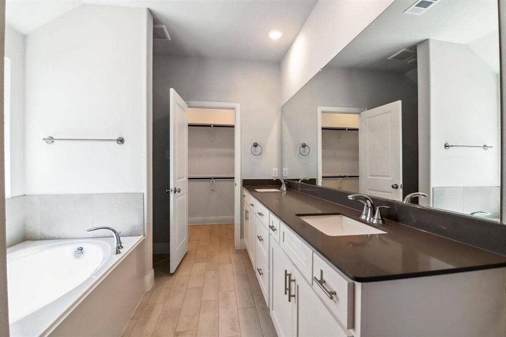 Dual vanity with integrated sinks, expansive wall-mounted mirror, built-in soaking tub with tile surround, wood-finish flooring, and a walk-in closet with shelving