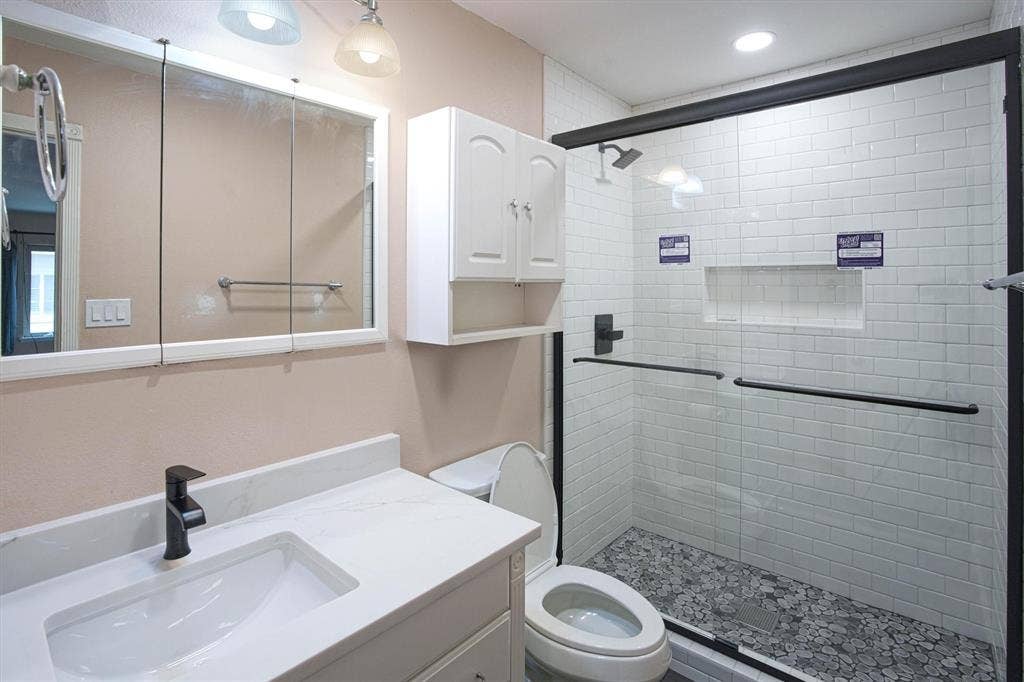 Bathroom featuring a vanity with a quartz counter top, integrated sink, matte black faucet, wall-mounted medicine cabinet, walk-in shower with white subway tile walls, and pebble-finish shower flooring