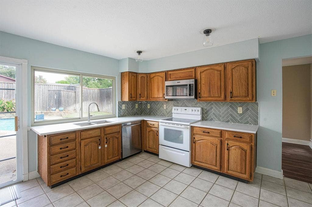 Kitchen featuring wood cabinetry, herringbone-pattern tile backsplash, stainless steel appliances, quartz countertops, and ceramic tile flooring