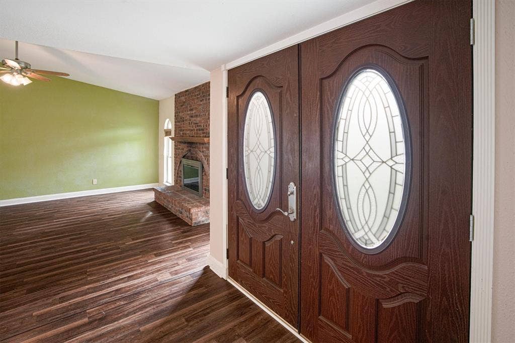 Double entry doors featuring decorative oval glass inserts, luxury vinyl plank flooring, vaulted ceilings, and a floor-to-ceiling brick fireplace with a wood mantel