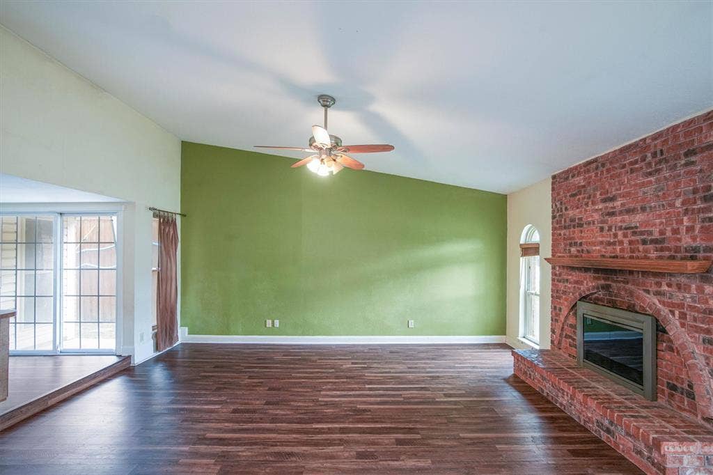 Vaulted ceiling featuring a central lighted ceiling fan, luxury vinyl plank flooring, exposed brick fireplace with a wood mantel, arched window, and large double paned sliding glass door