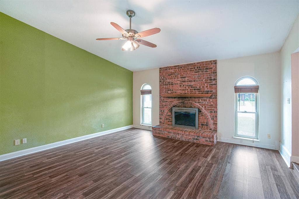 Vaulted ceiling featuring a ceiling fan, exposed brick fireplace with an arched hearth, wooden mantel, and luxury vinyl plank flooring with arched windows