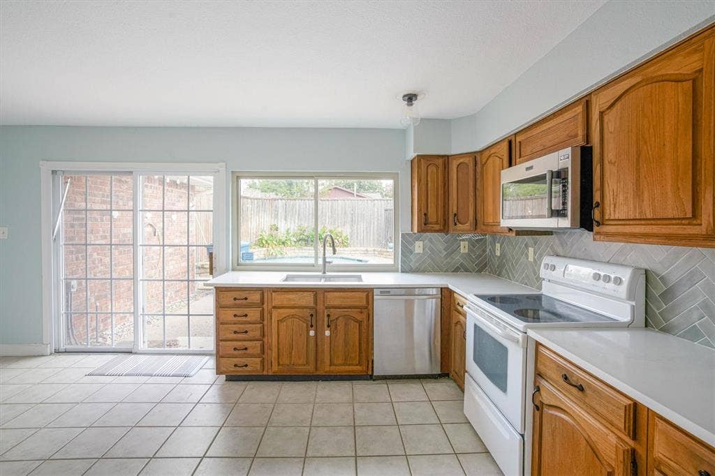 Kitchen featuring wood cabinetry, herringbone-pattern tile backsplash, quartz countertops, stainless steel appliances, and ceramic tile flooring