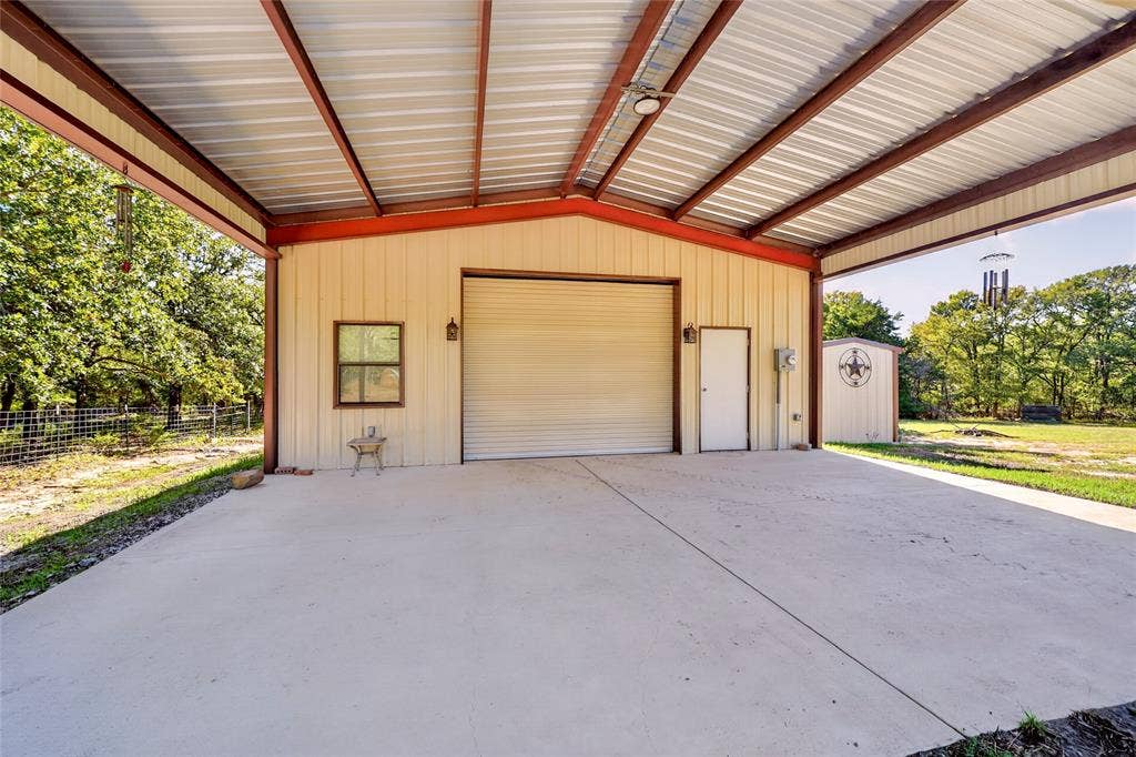 Expansive covered concrete pad with a metal overhead structure, featuring a roll-up garage door, an entry door, and an exterior window