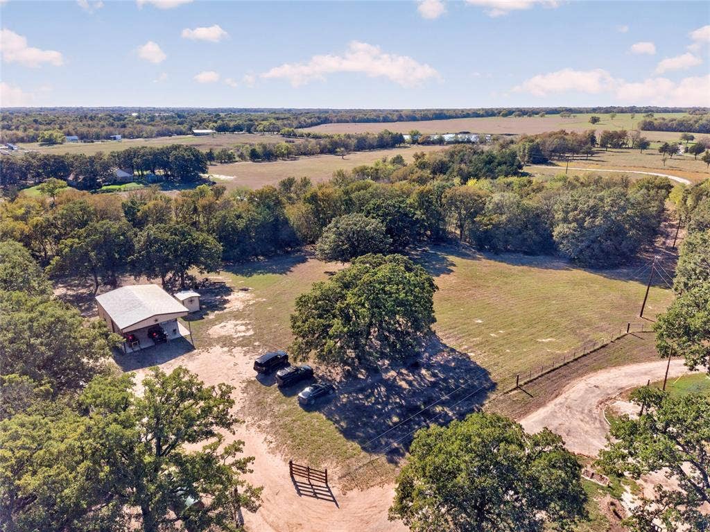 Expansive rural property featuring an outbuilding with a covered porch, mature trees, open grassy areas, a fenced perimeter, and a gravel driveway