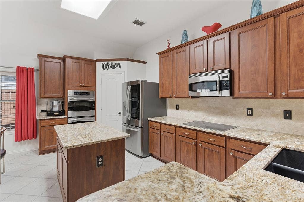 5104 Teddington Park Drive, Plano, TX 75023 Wood-finish cabinetry, light-toned stone countertops, a full suite of stainless steel appliances, a kitchen island, and a tile floor