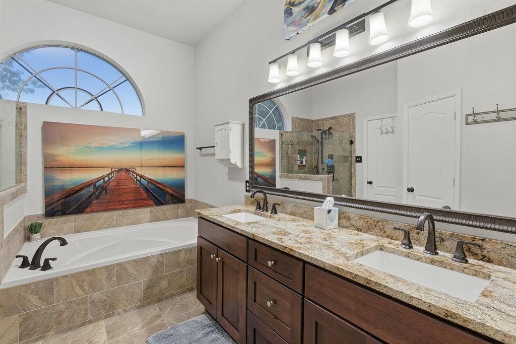 5104 Teddington Park Drive, Plano, TX 75023 Dual vanity with granite countertops and undermount sinks, large framed mirror with integrated lighting, built-in soaking tub with tiled surround, and arched window