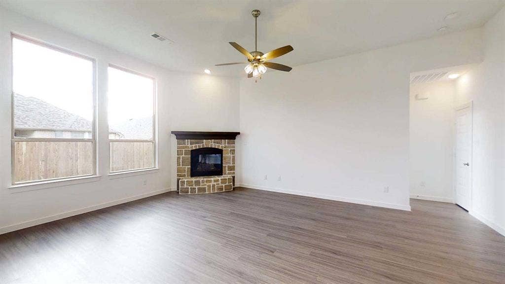 1553 Wyler Drive, Forney, TX 75126 Spacious room featuring wood-finish flooring, a corner fireplace with a stone surround and wood mantel, multiple windows, and a ceiling fan with integrated lighting
