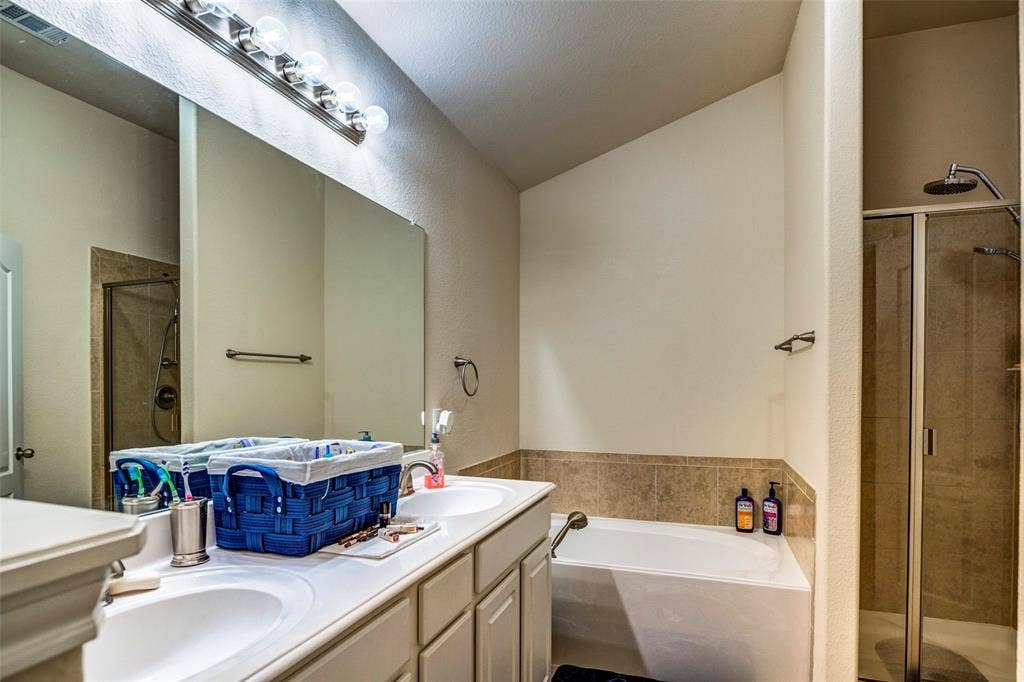 1040 Lucy Lane, Allen, TX 75013 Master bathroom featuring dual vanity with wood-finish cabinetry and white countertop, large framed mirror with integrated lighting, soaking tub with tile surround, and separate walk-in shower with glass enclosure and tile detailing