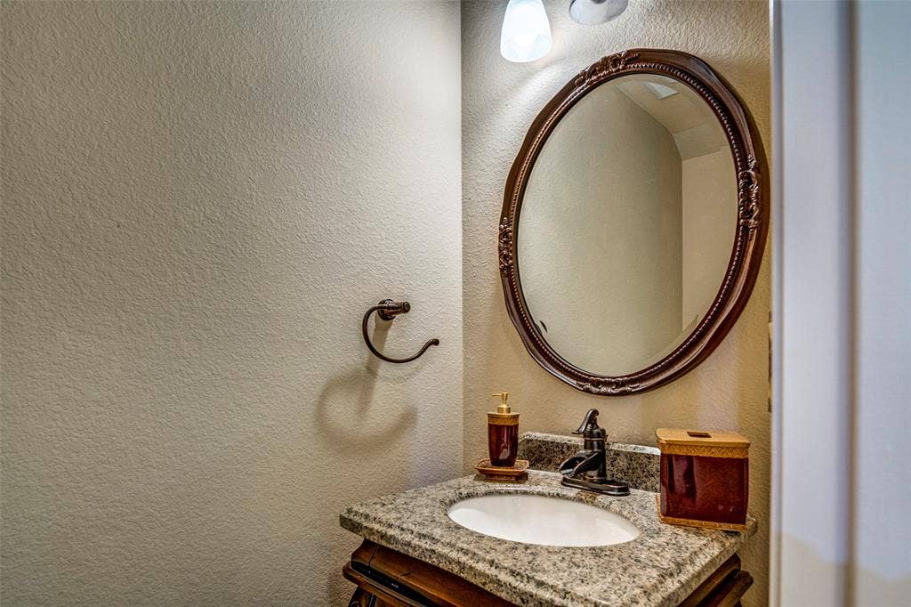 1040 Lucy Lane, Allen, TX 75013 Oval mirror with an ornate frame, granite countertop, undermount sink, and wall-mounted vanity lighting