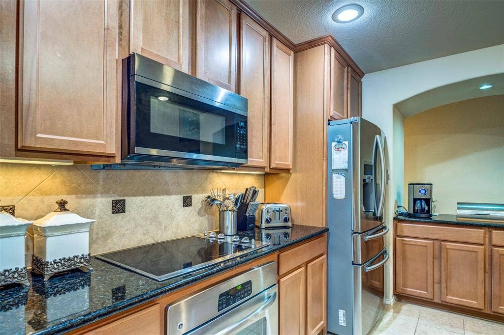 1040 Lucy Lane, Allen, TX 75013 Updated kitchen featuring warm wood cabinetry, black granite countertops, stainless steel appliances including a microwave oven and French-door refrigerator, and a decorative tile backsplash with recessed lighting throughout