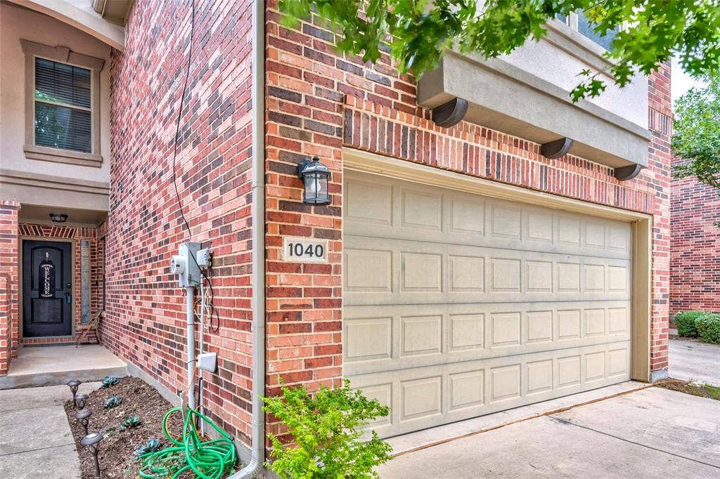 1040 Lucy Lane, Allen, TX 75013 Charming brick home at 1040 featuring a spacious two-car garage with paneled overhead door, manicured front landscaping with mature greenery, and classic red brick exterior with architectural detail