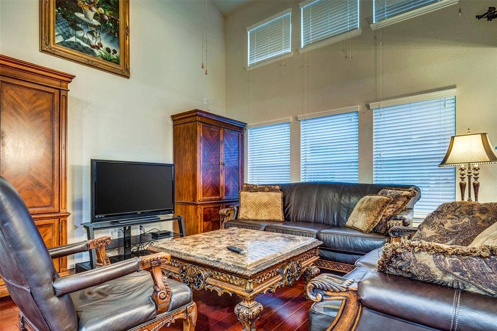 1040 Lucy Lane, Allen, TX 75013 Sophisticated living room showcasing soaring ceilings with transom windows, ornamental wood cabinetry with glass-front doors, elegant upholstered seating, decorative wood-finish flooring, and table lamps with brass accents