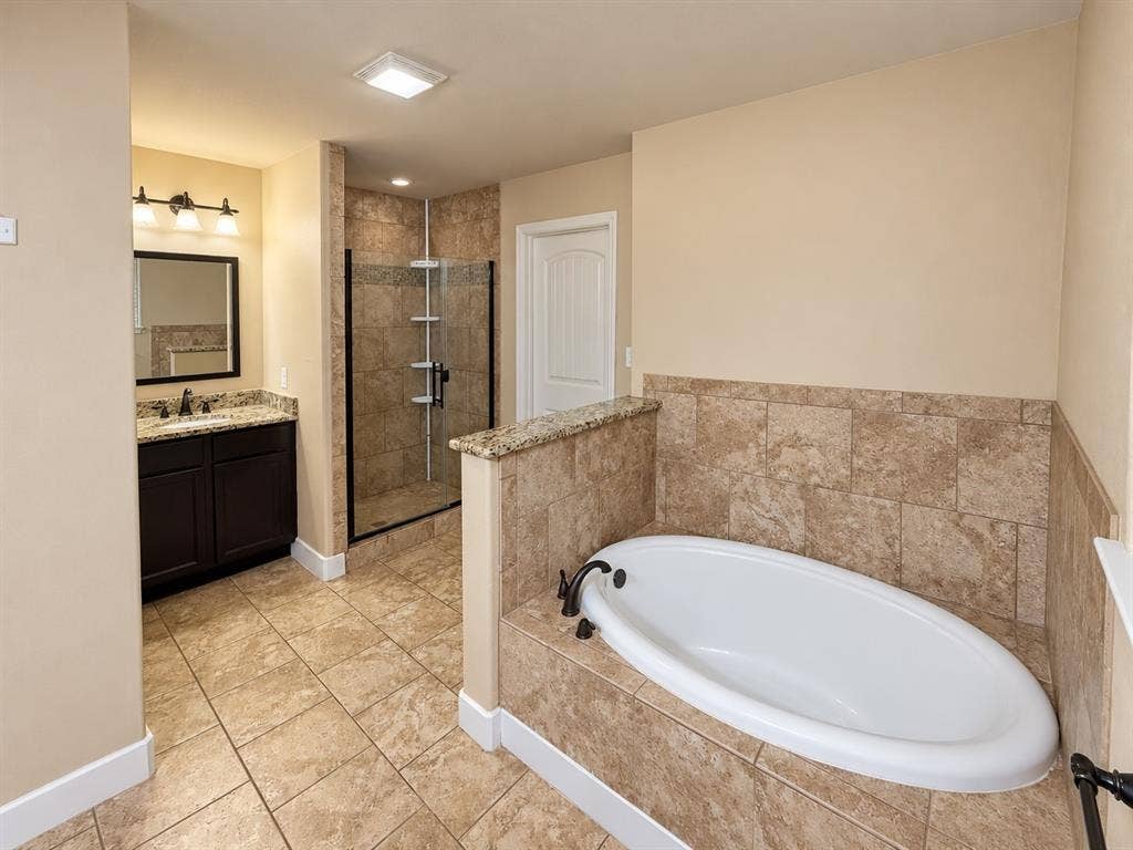 Bathroom featuring a tiled soaking tub, a separate glass-enclosed shower with tiled walls, a vanity with a stone countertop, and tiled flooring throughout