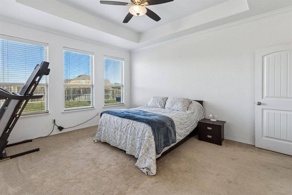 1501 Coyote Ridge Road, Wylie, TX 75098 Tray ceiling featuring a ceiling fan, three windows with horizontal blinds, carpeted flooring, and a paneled door with an arched top