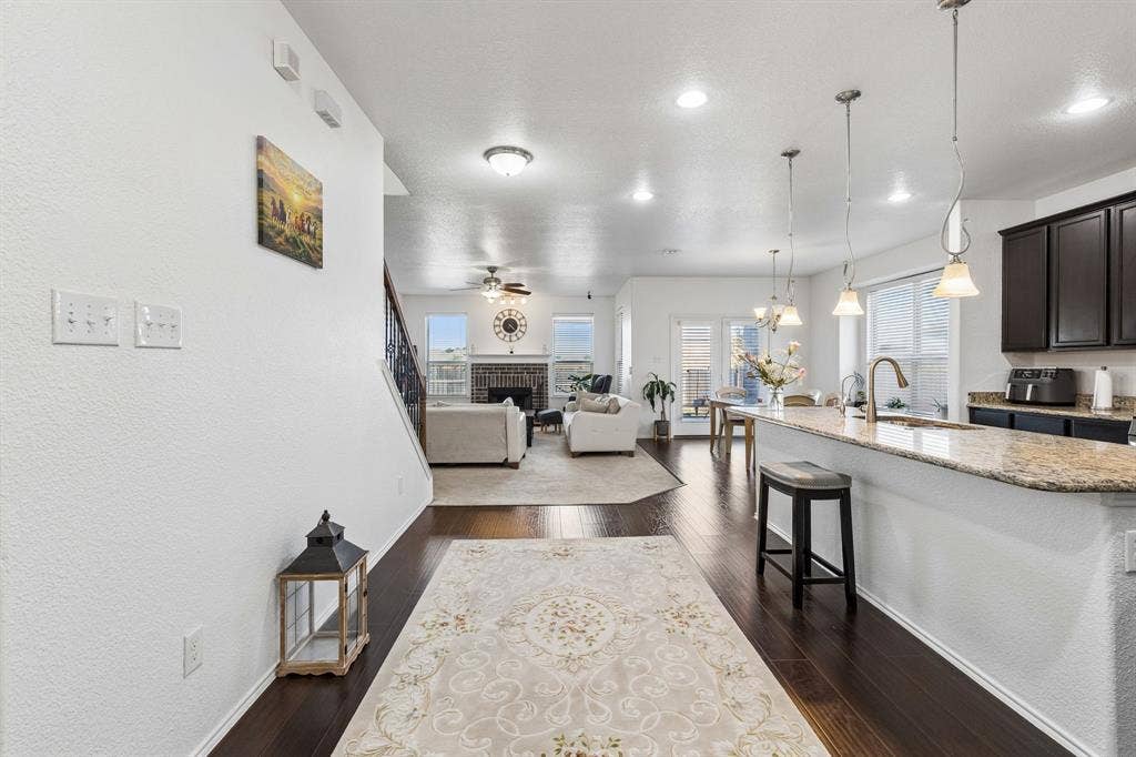 1501 Coyote Ridge Road, Wylie, TX 75098 Dark wood-finish flooring extending into the living area, kitchen island with granite countertops and pendant lighting, dark wood cabinetry, and a living area featuring a fireplace with a stone surround