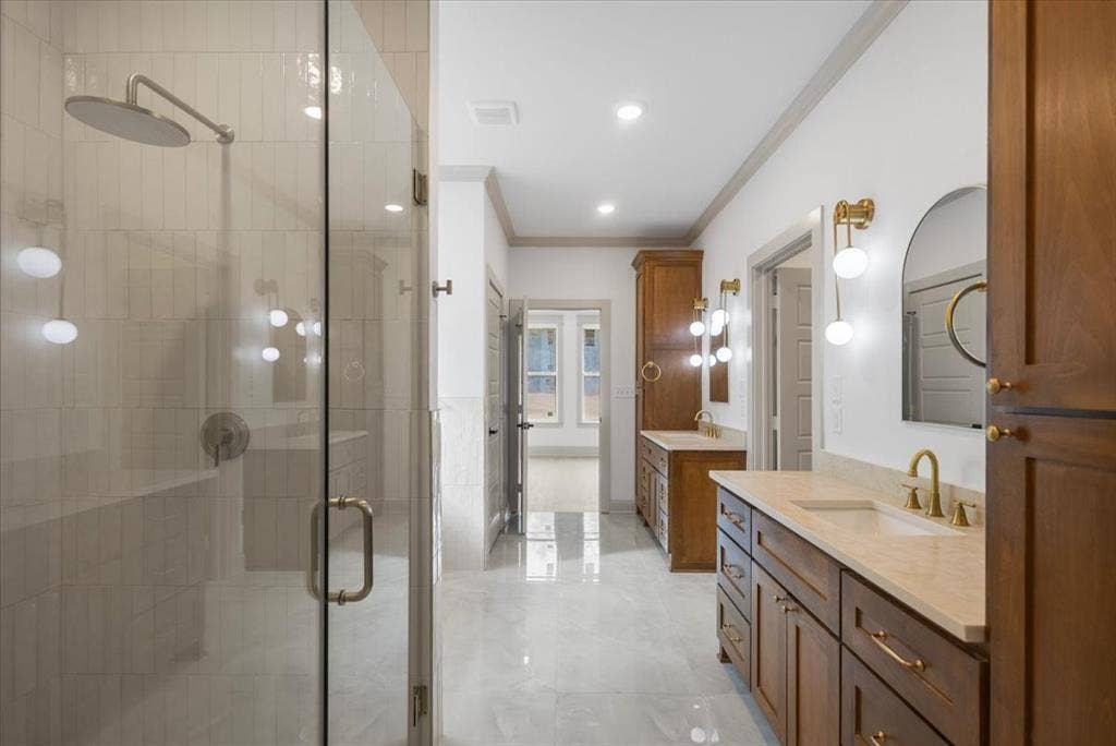 17155 Island Lake Dr., Tyler, TX 75708 Spacious bathroom featuring a glass-enclosed shower with a rainfall showerhead, dual wood-finish vanities with integrated sinks and light-colored countertops, and gold-finish hardware throughout