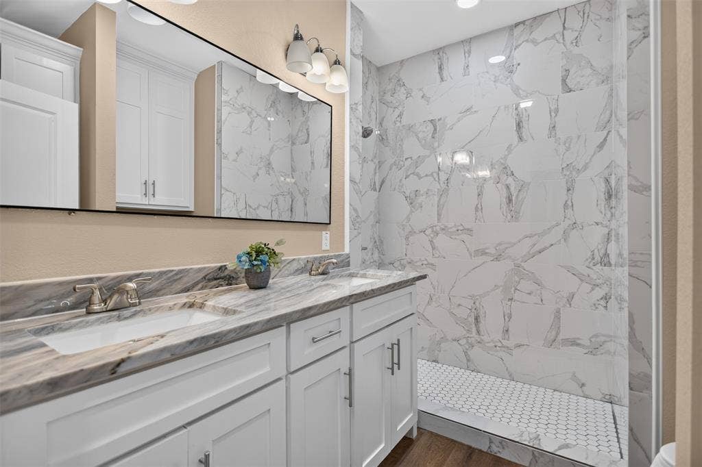 Visitor Bathroom featuring a dual-sink vanity with stone-surface countertop, white cabinetry with silver hardware, full-width mirror, and a walk-in shower with floor-to-ceiling decorative tile and a mosaic tile floor
