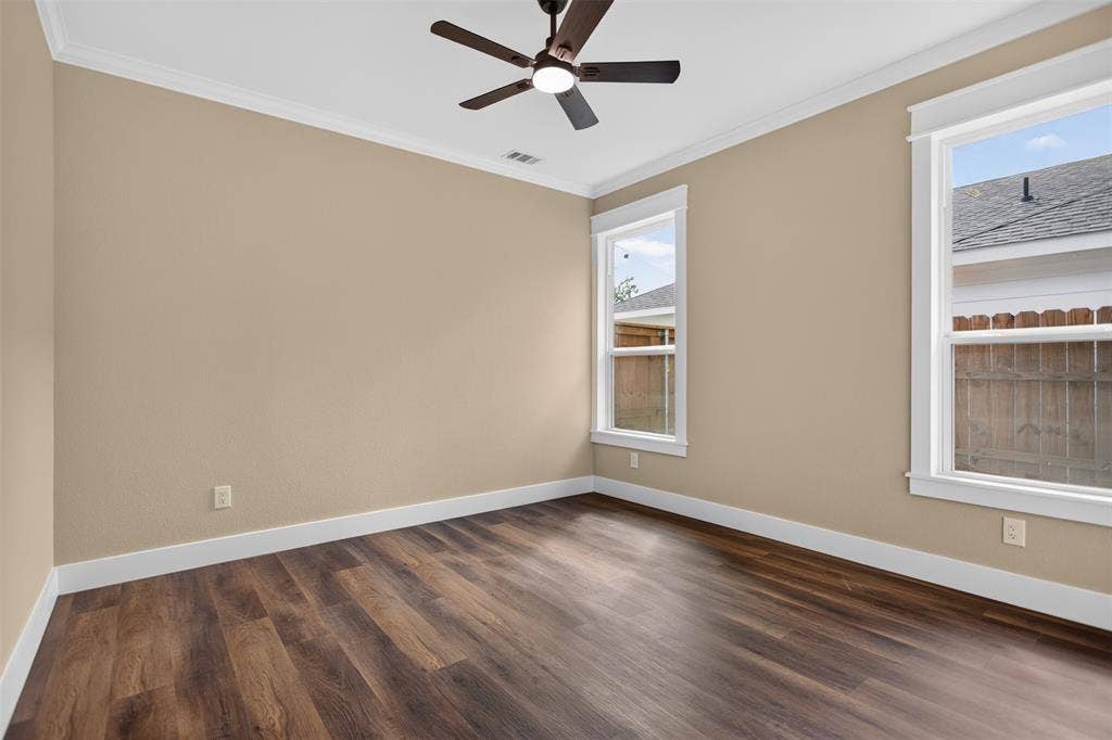 Neutral-toned interior space featuring wood-finish flooring, dual windows with white trim, crown molding, and a ceiling-mounted fan with light fixture