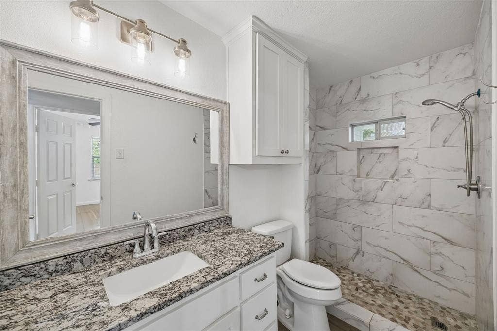 2245 County Road 337, McKinney, TX 75071 Bathroom featuring a vanity with a stone countertop, an integrated rectangular sink, wall-mounted light fixtures, built-in cabinetry, and a walk-in shower with stone-patterned wall tile and a pebble-stone floor
