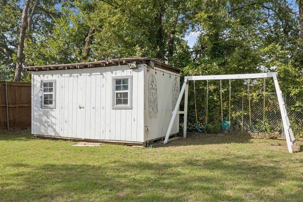 2245 County Road 337, McKinney, TX 75071 Outdoor storage shed featuring vertical siding, two windows, and integrated exterior lighting, situated on a grass lot with perimeter wood and chain-link fencing