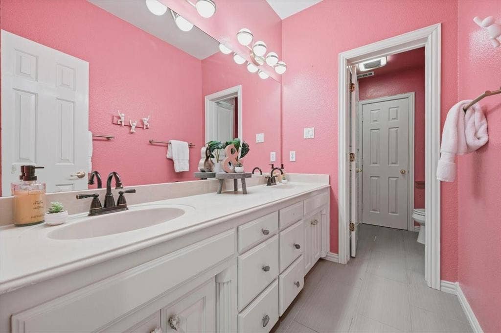 2945 Emerald Sound Drive, Cedar Hill, TX 75104 Bathroom featuring a dual-sink vanity with a solid-surface countertop, dark metal hardware, a full-width wall mirror, and a multi-bulb light fixture