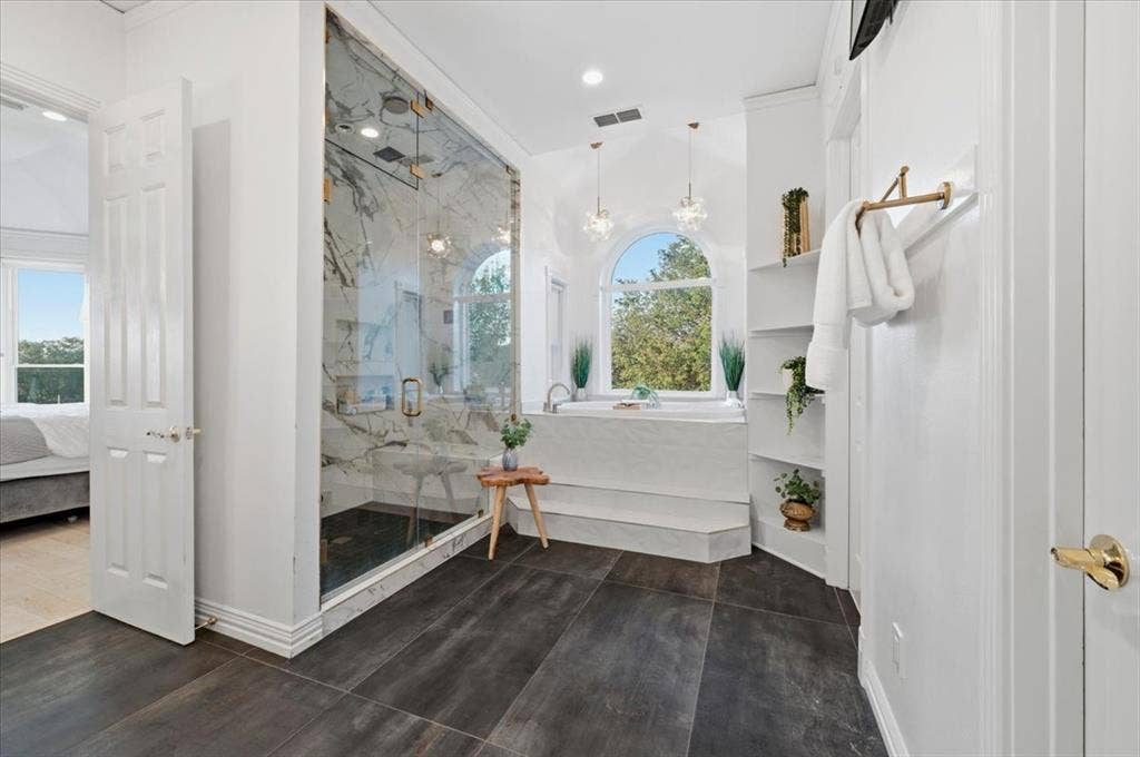 2945 Emerald Sound Drive, Cedar Hill, TX 75104 Walk-in shower with stone-finish tile surround and glass enclosure, deep soaking tub, arched window, and built-in shelving, complemented by dark large-format tile flooring