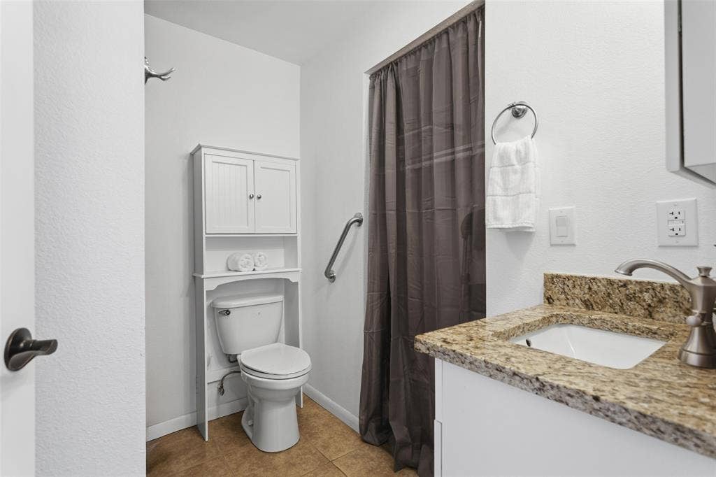 Granite-finish countertop with an undermount sink, built-in white cabinetry, tiled flooring, and a shower with a grab bar