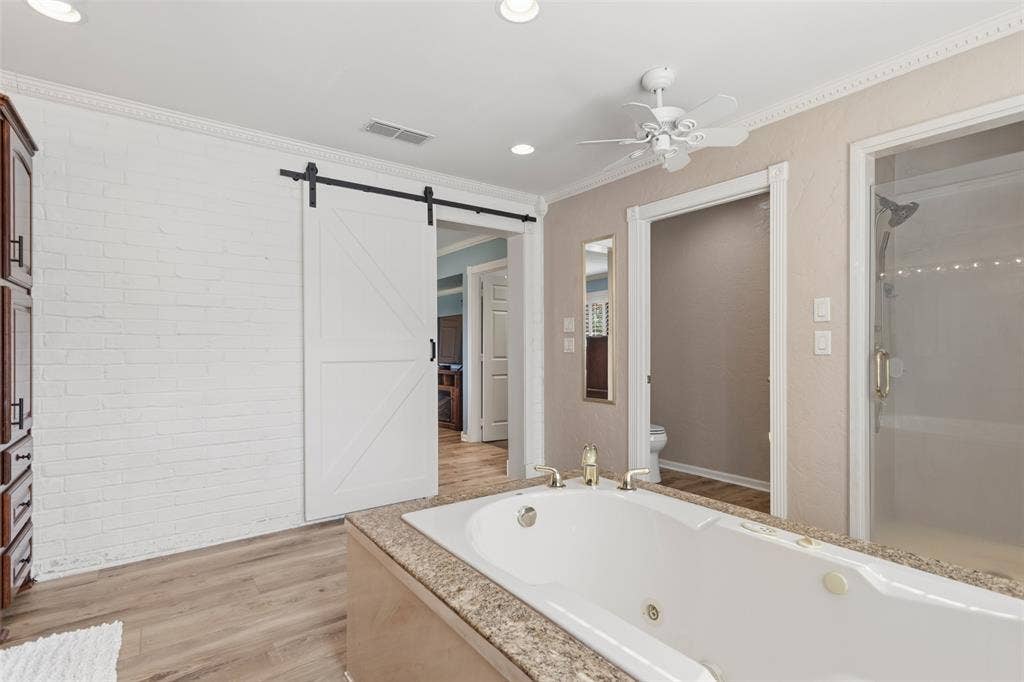 Soaking tub with a speckled countertop, walk-in shower with clear glass enclosure, ceiling fan, and wood-finish flooring