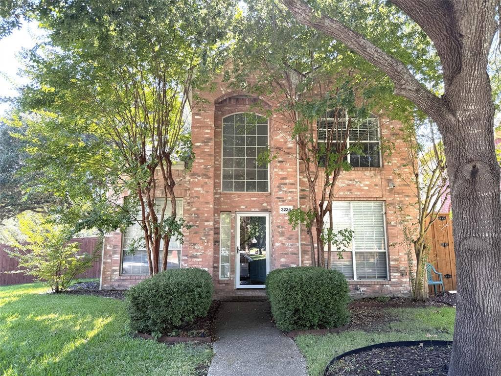 Brick exterior featuring an arched window, large picture windows, mature trees, manicured lawn, and a concrete pathway