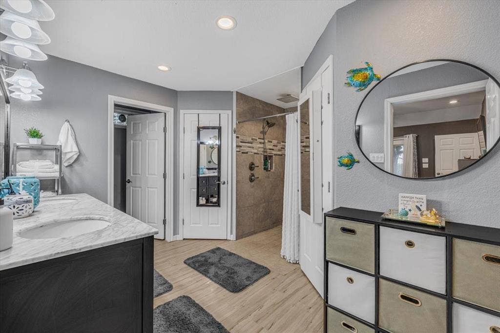 1106 Scenic Drive, Granbury, TX 76048 Bathroom featuring stone-finish vanity countertops with dual recessed sinks, dark cabinetry, wood-finish flooring, and a walk-in shower with tiled surround and an integrated niche