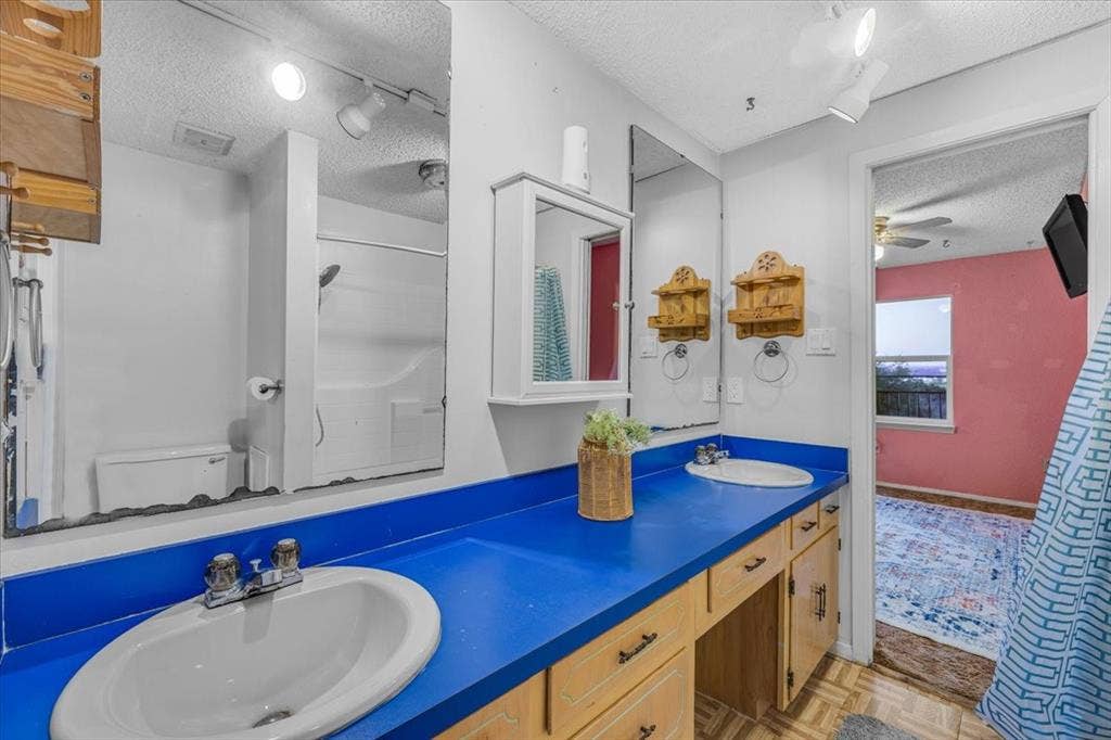 1106 Scenic Drive, Granbury, TX 76048 Bathroom vanity featuring an extended solid-surface countertop, dual integrated oval sinks, light-wood cabinetry, a wall-mounted mirrored medicine cabinet, and large-format wall mirrors