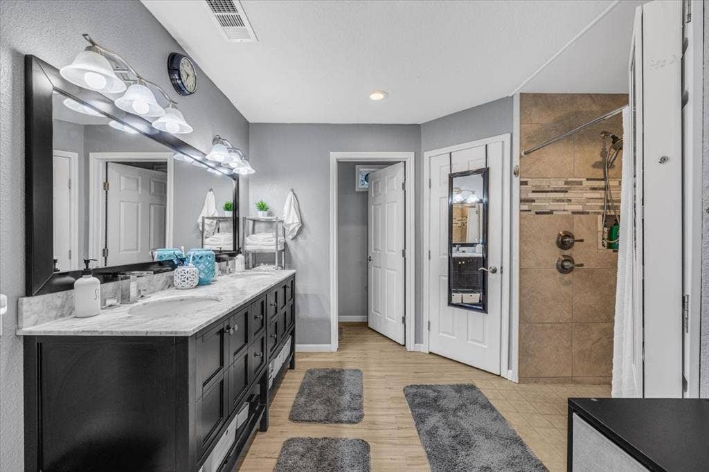 1106 Scenic Drive, Granbury, TX 76048 Double-sink vanity featuring a stone-surface countertop, dark-finish cabinetry, a full-width mirror, integrated wall-mounted lighting, and a walk-in shower with decorative tile inlay