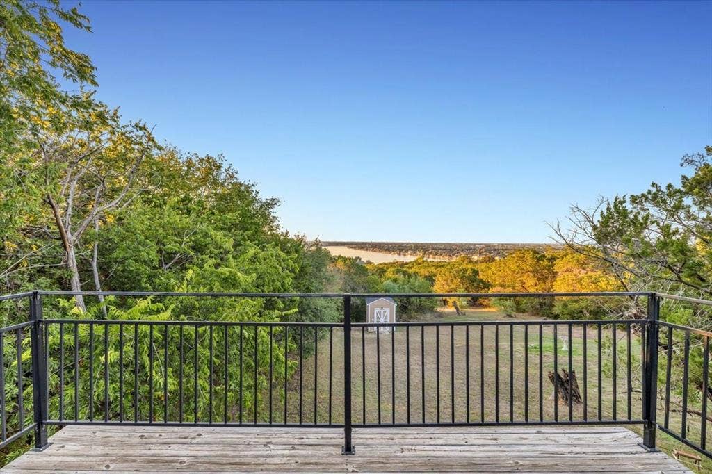 1106 Scenic Drive, Granbury, TX 76048 Elevated wooden deck with black metal railing, overlooking a fenced outdoor area featuring mature trees and a detached storage structure