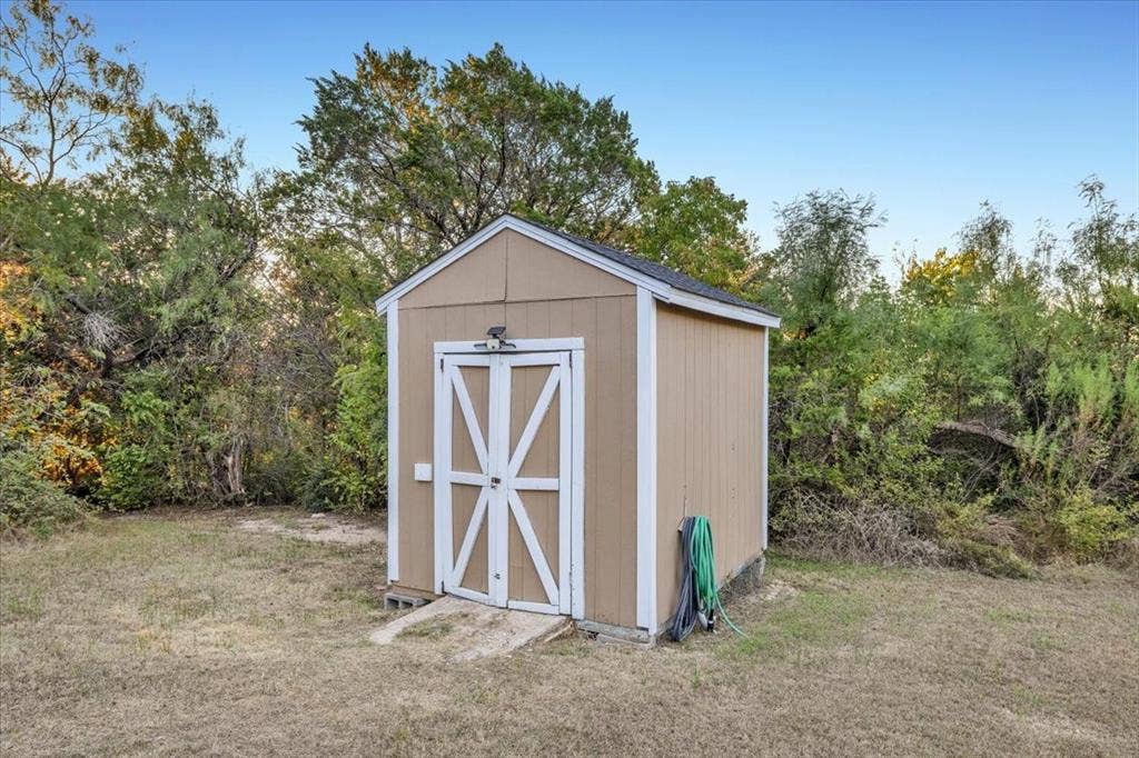 1106 Scenic Drive, Granbury, TX 76048 Storage shed featuring wood-panel siding, double doors with X-bracing, a shingled roof, and a concrete entry pad