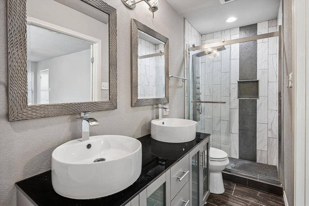 Dual vessel sinks set on a black countertop, glass-enclosed shower with contrasting tile, wood-finish flooring, and wall-mounted vanity lighting