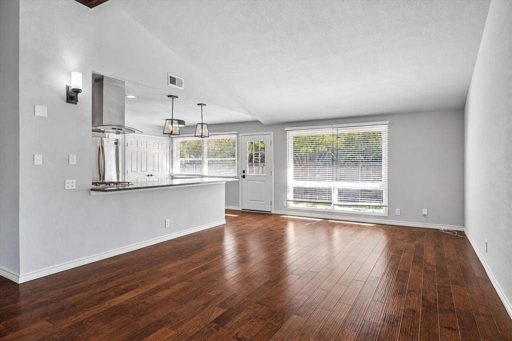 Rich wood-finish flooring, vaulted ceiling, white cabinetry with dark hardware, stainless steel appliances, and large windows with privacy blinds