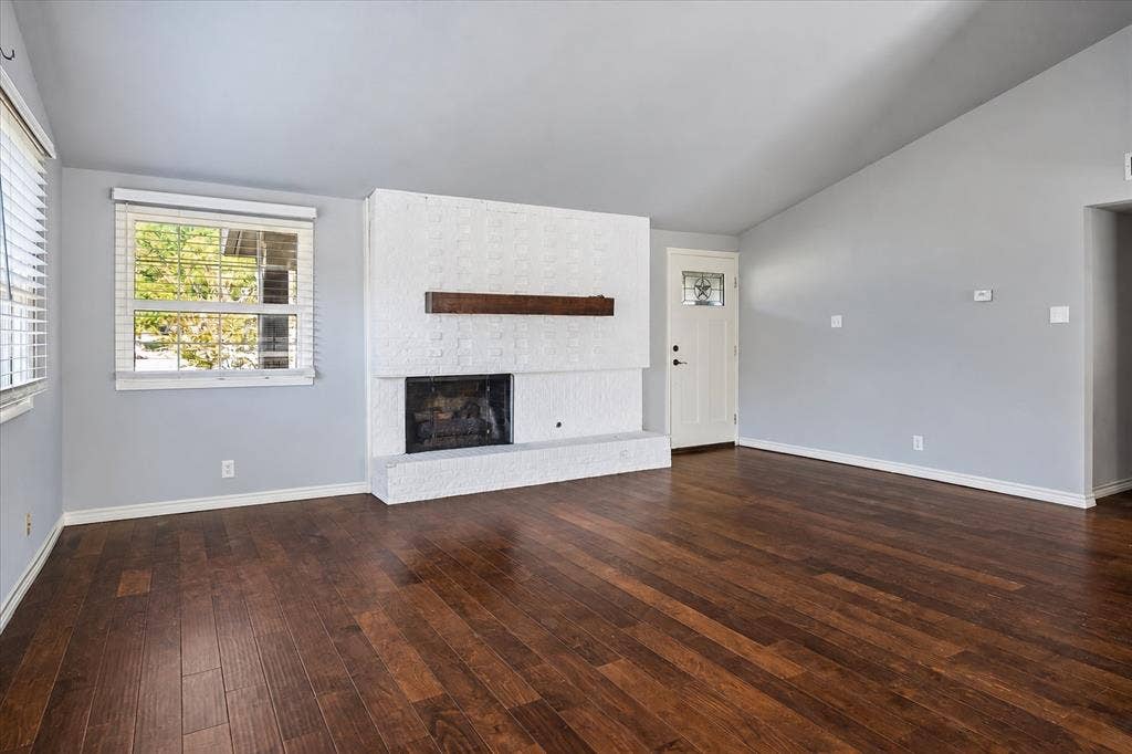 Vaulted ceiling, wood-finish flooring, white brick fireplace with wood mantel, white entry door with decorative glass inlay, and light gray painted walls