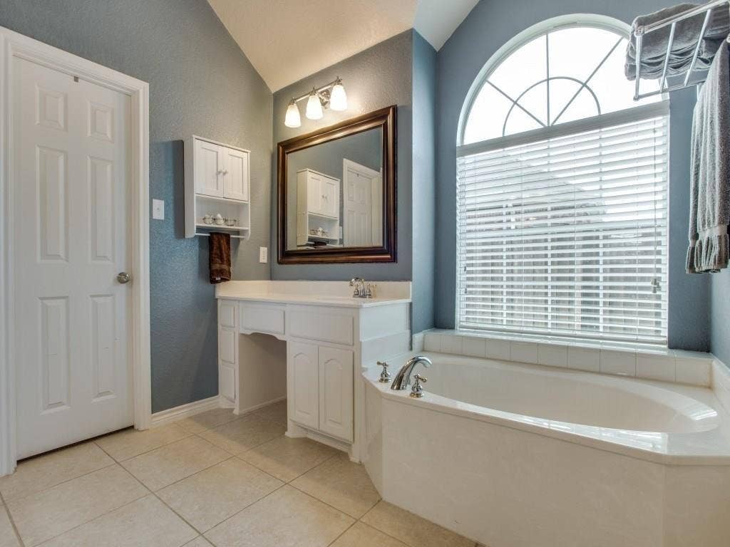 12316 Sailmaker Lane, Frisco, TX 75035 Vaulted ceiling, arched window with grilles, built-in bathtub with deck, white vanity with integrated sink, and large format tile flooring