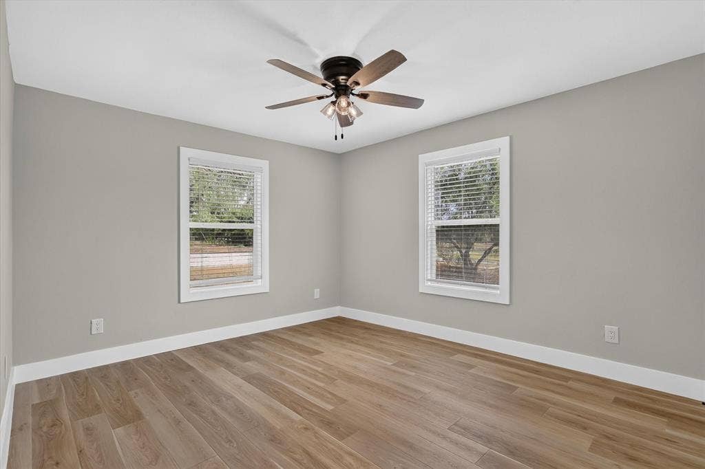 Wood-finish flooring, light gray walls, two windows with white trim, and a ceiling fan with integrated lighting