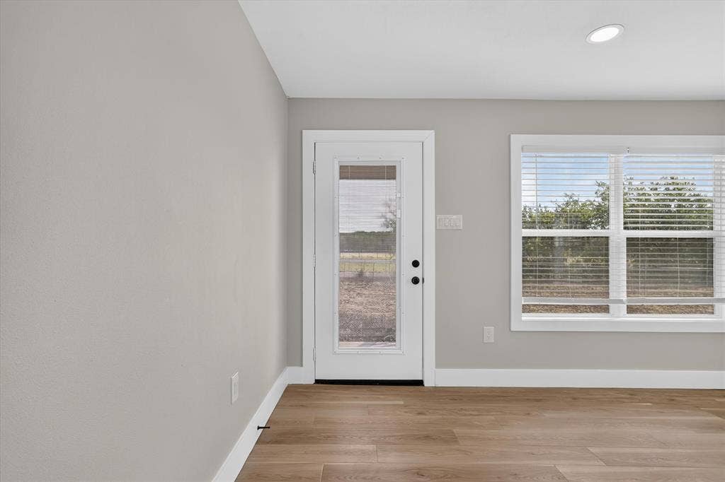 Bright and airy room showcasing gleaming wood-finish flooring, white trim detailing, and abundant natural light from a full-glass entry door and large dual-pane windows with integrated blinds