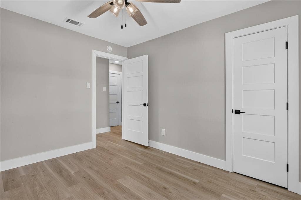 Wood-finish flooring, contemporary light grey wall color, modern ceiling fan with integrated lighting, white five-panel interior doors, and black door hardware