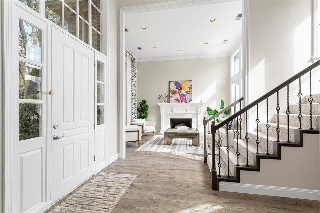A striking glass-paneled front door welcomes you into a bright foyer featuring beautiful hardwood-style flooring, a sweeping staircase with iron balusters, and a clear view into the formal living room.
