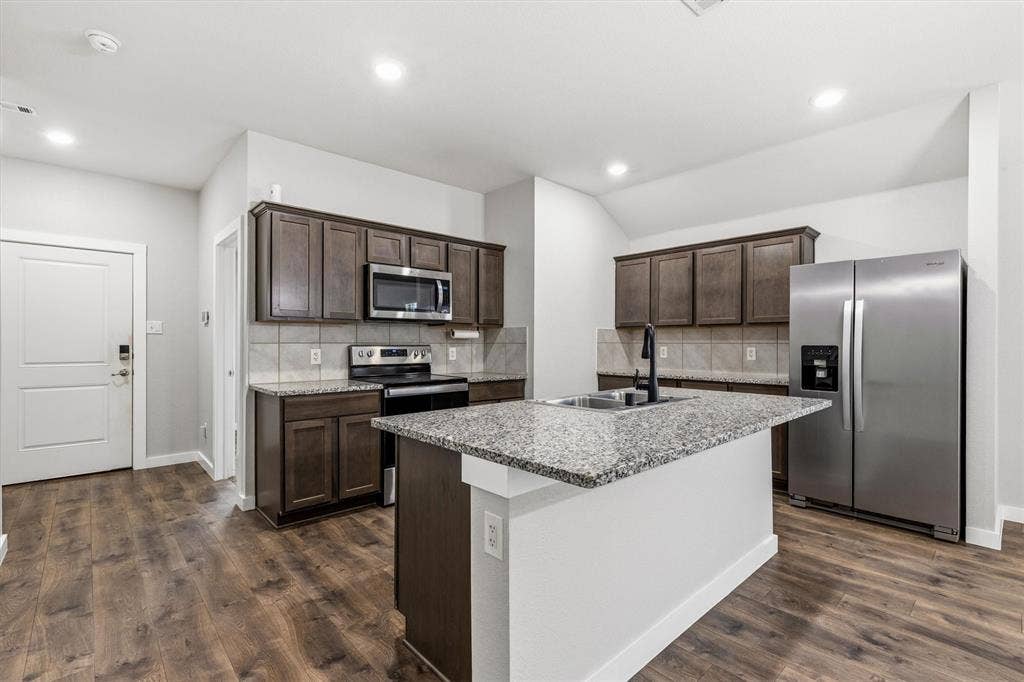2333 Bellewood Court, Crandall, TX 75114 Kitchen featuring wood-finish flooring, dark wood cabinetry, granite countertops, an island with an undermount sink, and stainless steel appliances
