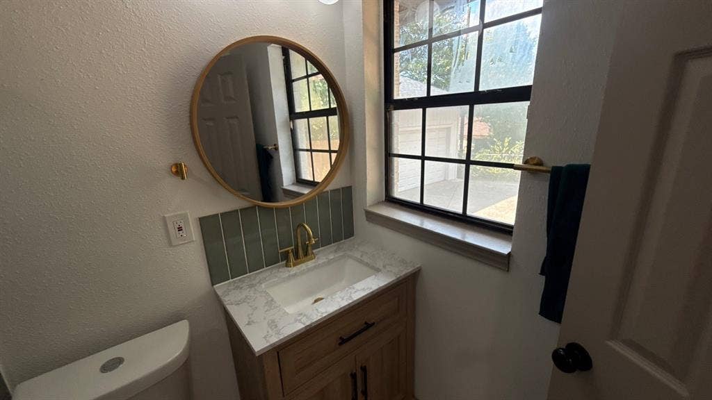 7437 Eccles Drive, Dallas, TX 75227 Wood-finish vanity with a stone-patterned countertop, integrated basin, vertical tile backsplash, round mirror, and brass-finish hardware