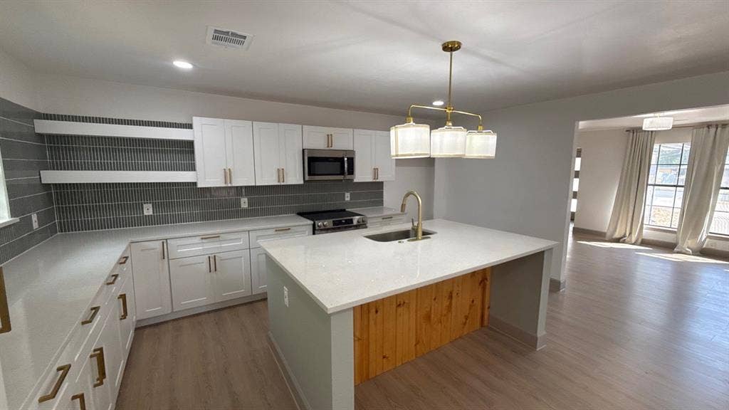 7437 Eccles Drive, Dallas, TX 75227 Kitchen featuring white shaker-style cabinetry, a central island with wood-paneling detail, quartz countertops, a linear tile backsplash, and metallic hardware