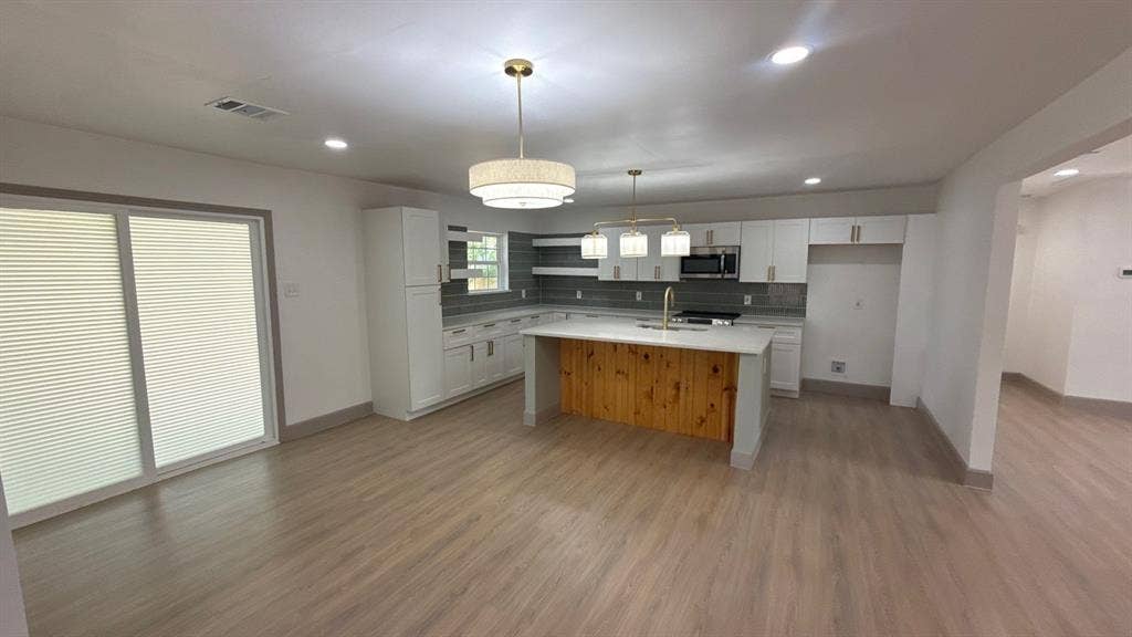 7437 Eccles Drive, Dallas, TX 75227 Kitchen featuring wood-finish flooring, a central island with a wood-paneled base and solid surface countertop, white shaker-style cabinetry, a tile backsplash, and recessed and pendant lighting