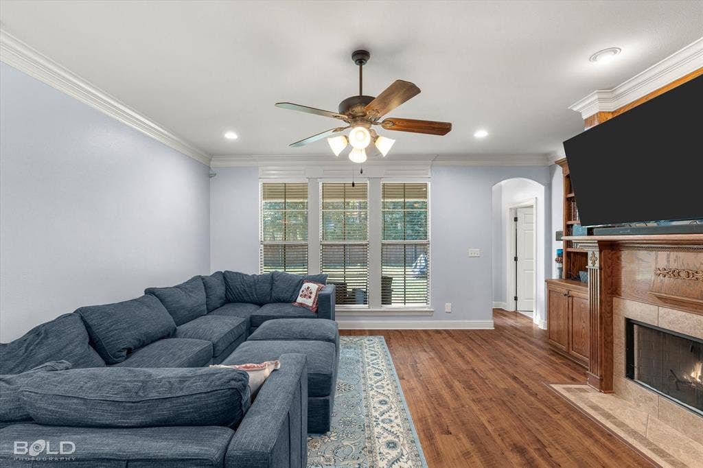 Wood-finish flooring, custom built-in wooden cabinetry, fireplace with a stone-surround, crown molding, and ceiling fan with integrated lighting