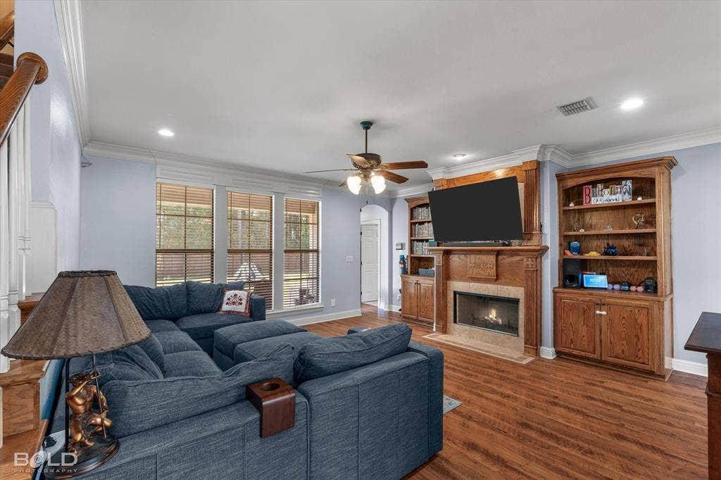 Living area featuring wood-finish flooring, a wood-surround fireplace with a tiled hearth, integrated custom wooden cabinetry, crown molding, and large multi-pane windows